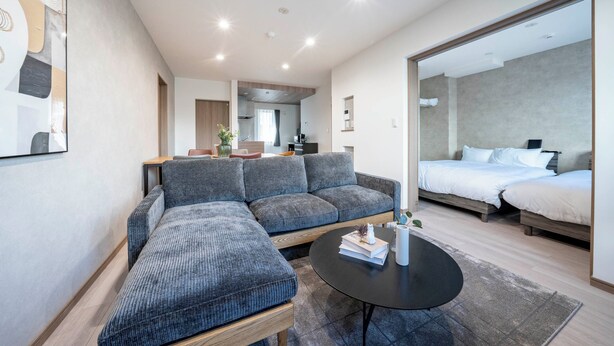 Ein modernes Hotelzimmer mit einer grauen Eckcouch, einem kleinen runden Tisch, einem Bett mit weißen Bettwäsche und einem großen Spiegel.