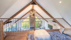 Une chambre à coucher aménagée dans un espace mansardé, avec un plafond en pente, des poutres en bois, une grande fenêtre, un luminaire suspendu et une vue sur l’extérieur.