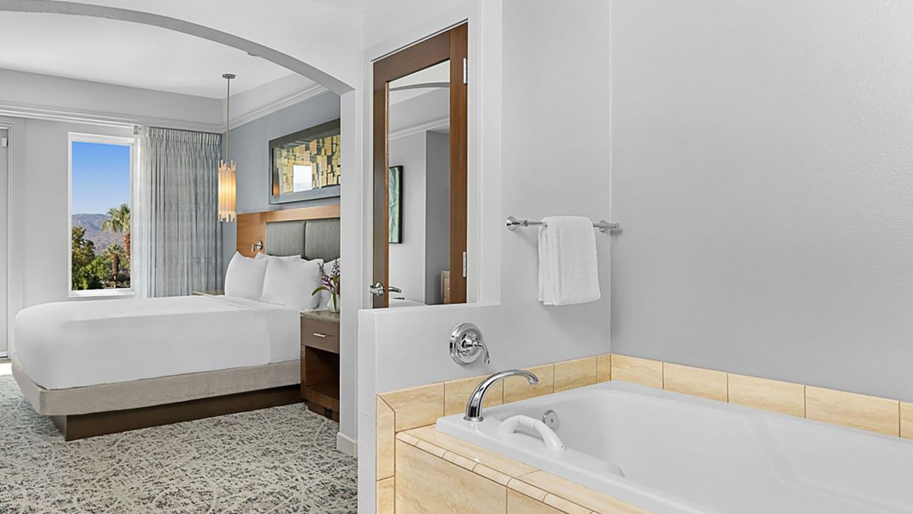 A hotel room with a large bed, a separate bathroom with a bathtub, a large mirror, and a view of mountains through a window.