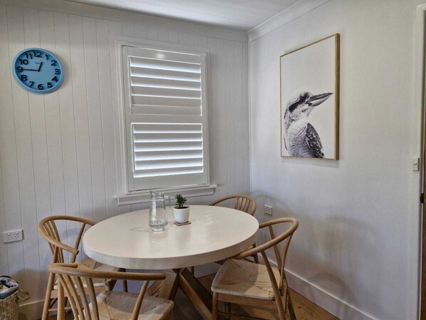 A dining area with a round table, four wooden chairs, a wall clock, a window with blinds, and a framed bird artwork. - Caves Beach Coastal Getaway - Perfect for Families (Caves Beach)