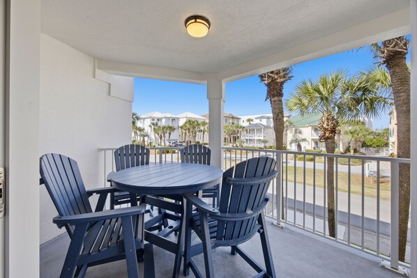 A covered outdoor seating area with a table and chairs, a railing, and a view of palm trees and buildings in the background. - New Owner, New Look! Maravilla 3203 - 2 bed/2 Bath with Beach Service for 4! (Miramar Beach)