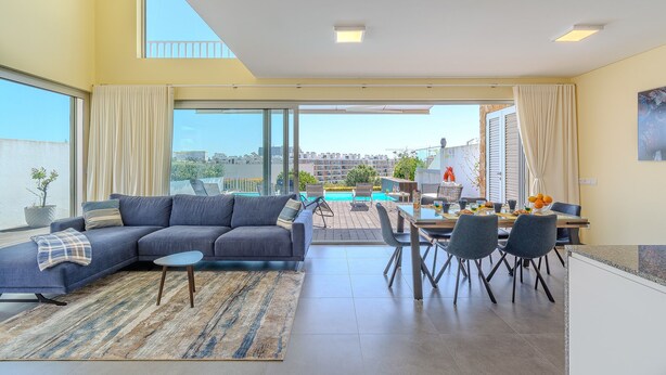 A modern living room with a sofa, dining table, and chairs. There is a large window with curtains, a view of a pool and buildings outside. - Villa Aleksandra - Private Pool (Albufeira)