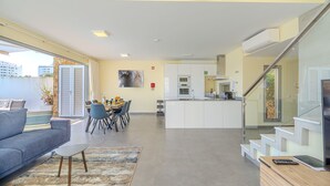 A modern living room with a sofa, dining table, and chairs, a kitchen with white cabinets, and a staircase. - Villa Aleksandra - Private Pool (Albufeira)