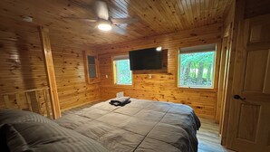Une chambre de style cabane en bois, avec deux lits, un ventilateur de plafond, un téléviseur fixé au mur et des fenêtres donnant sur la forêt.
