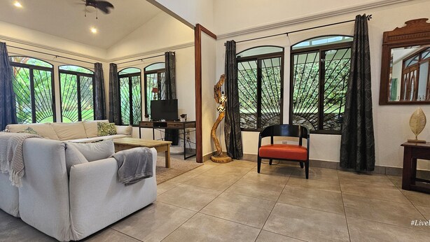 A spacious living room with a sofa, a coffee table, and a TV. There are large windows with curtains, a ceiling fan, and a decorative item on a side table. - Casa Suryaa Ananda in the heart of Ojochal (Provincia de Puntarenas)