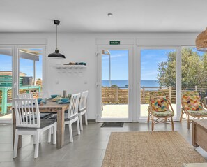 A modern dining area with white chairs, a wooden table, and a large window offering a view of the sea. - Family cottage in Benajarafe w pool and sea view (Vélez-Málaga)