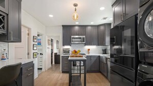 A modern kitchen with dark cabinetry, a central island, stainless steel appliances, and a hanging light fixture.