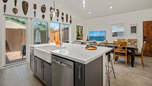 A modern kitchen with a large island, dining area with wooden chairs, and a view of the sea through large windows. - NEW Rustic Villa – 2BD, 2BA Close to Beach and Everything! (Santa Barbara)