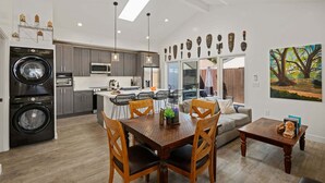 A modern kitchen with a built-in washer and dryer, a dining area with a wooden table and chairs, and a living room with a sofa and a painting. - NEW Rustic Villa – 2BD, 2BA Close to Beach and Everything! (Santa Barbara)