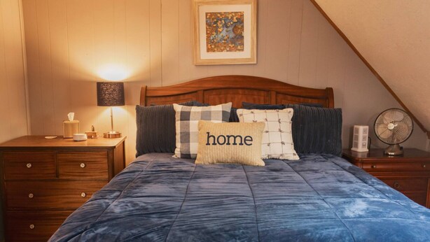 A bedroom with a wooden headboard, a blue quilted bedspread, and a 'home' pillow. There is a bedside table with a lamp, a small bedside table with a tissue box, and a fan on the right. - Aspen Room Serene Retreat at Columbine (Silverton)