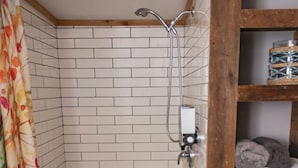 A small bathroom with white tiled walls, a shower, a wooden stool, and a shelf with a basket. - Aspen Room Serene Retreat at Columbine (Silverton)