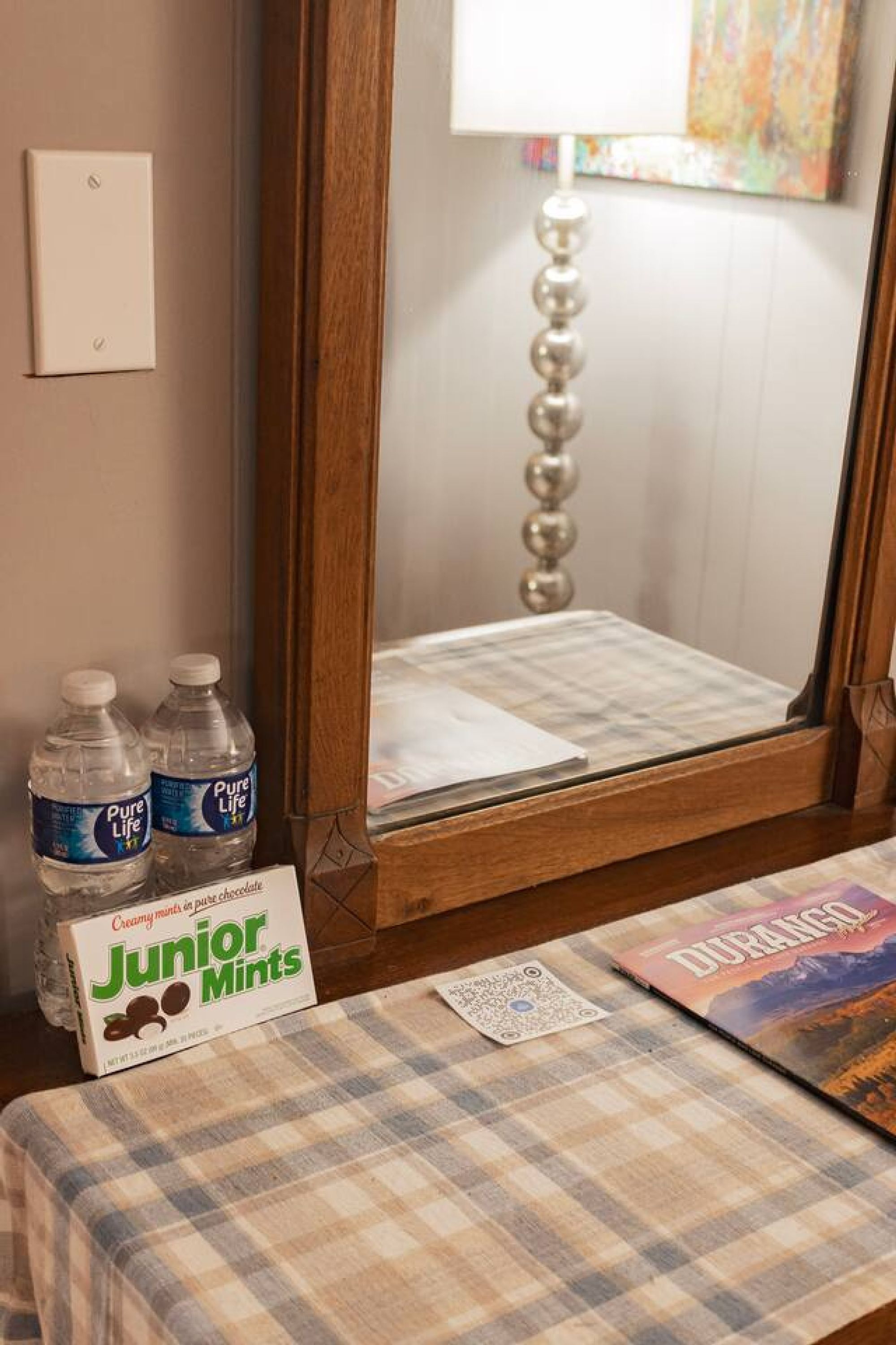 Una habitación con un espejo de madera, un mantel a cuadros, dos botellas de agua Pure Life, una caja de Junior Mints, una carta de juego y un libro titulado 'DURANGO'.