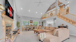 A spacious living room with a large beige sofa, a fireplace, a dining area with wooden furniture, and a staircase leading to an upper level. - Large Family Friendly House on the Fairway Hideout (Lake Ariel)