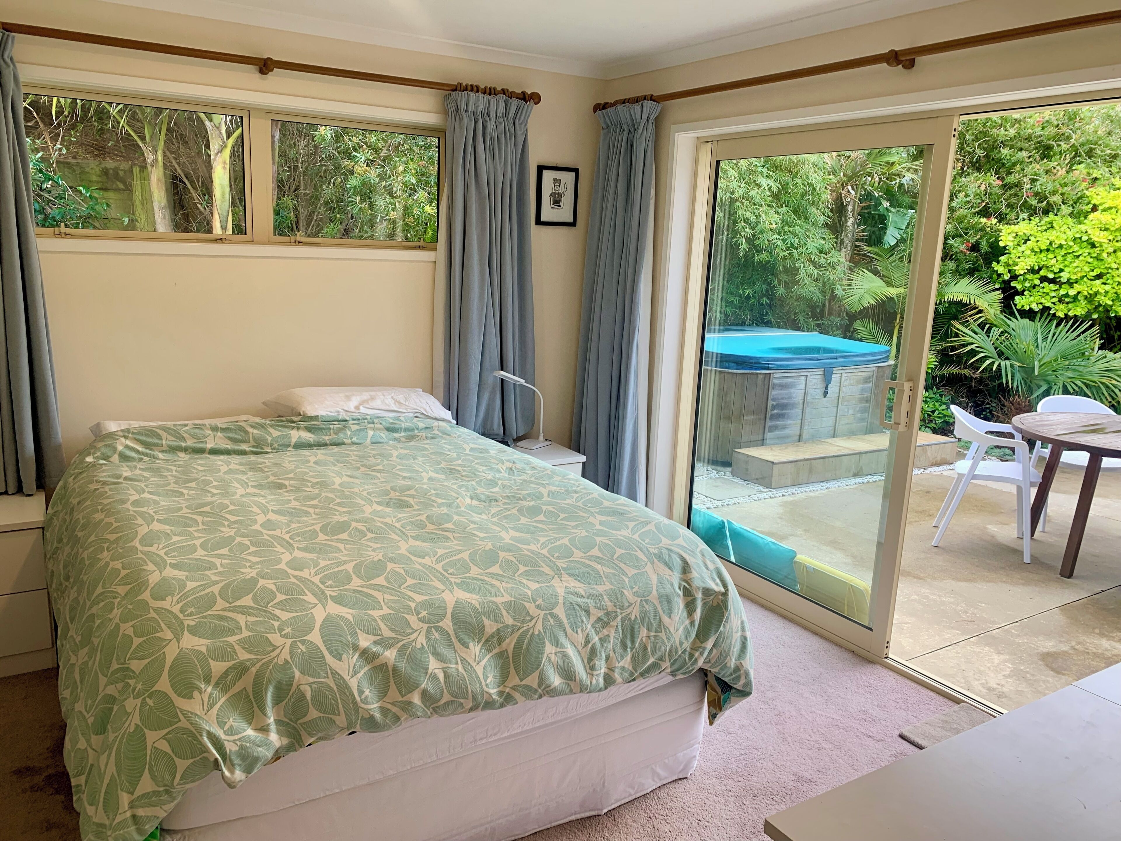 A bedroom with a bed, a bedside table, a window with curtains, and a view of a garden and pool outside.