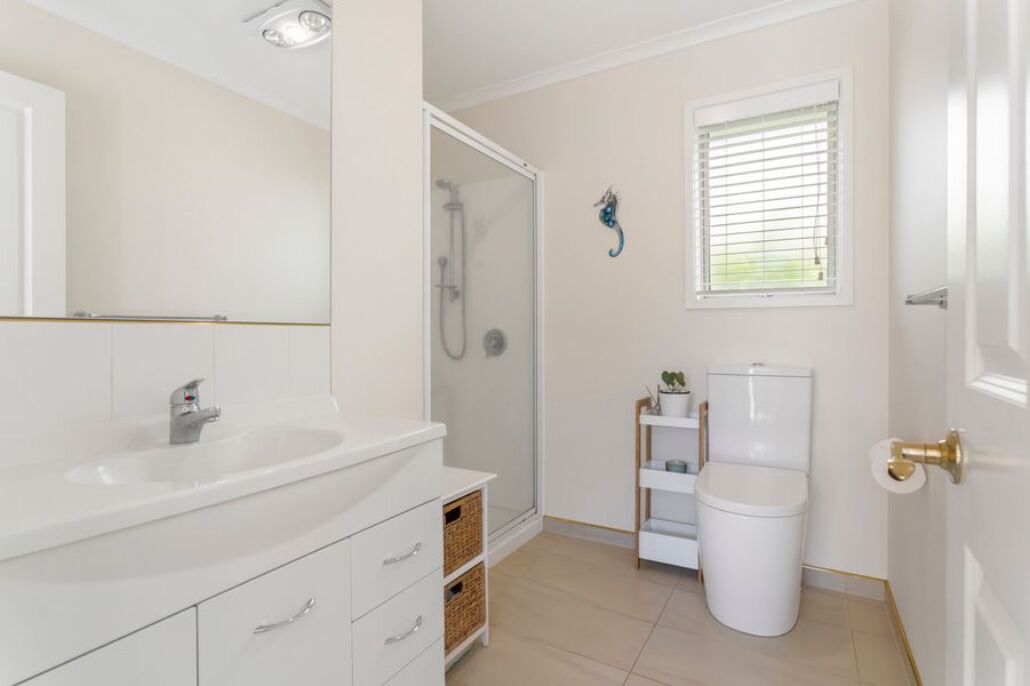 Ein Badezimmer mit weißem Waschbecken, einer Duschkabine aus Glas, einer Toilette und einem Fenster mit Jalousien.