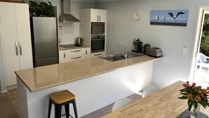 A modern kitchen with a central island, white cabinetry, stainless steel appliances, and a dining area with a wooden table and chairs. - Private Coastal Sanctuary, Next to the beach! (Coastlands)