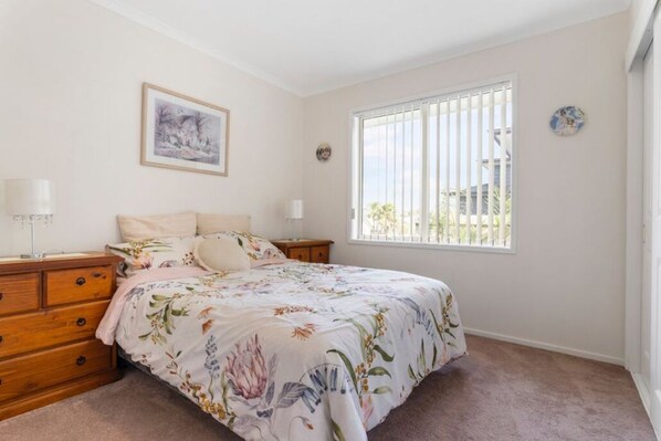 A bedroom with a floral bedspread, a wooden dresser, two bedside tables with lamps, a window with blinds, and a framed picture on the wall.