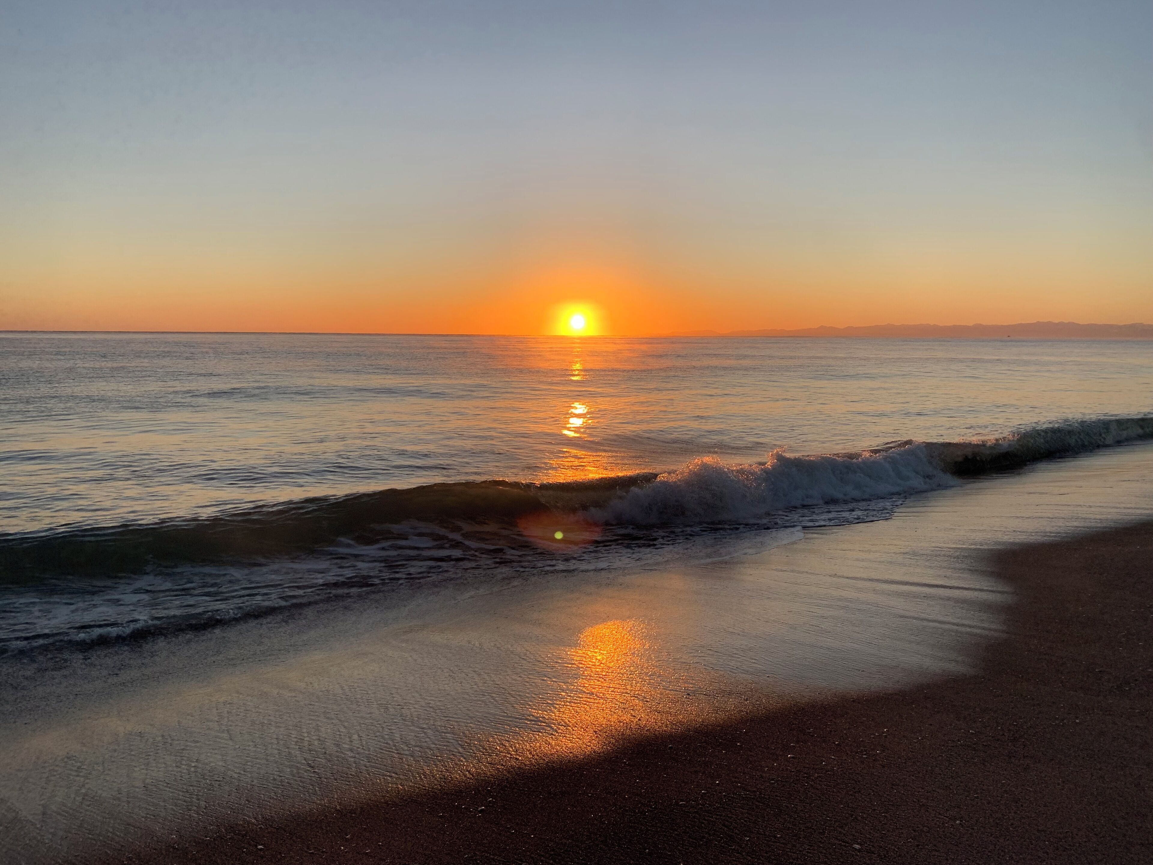 Un coucher de soleil sur une plage, avec des vagues qui viennent doucement lécher le rivage.