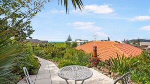 A patio with a round table and two chairs, surrounded by lush greenery and a view of a town with red-roofed buildings. - Private Coastal Sanctuary, Next to the beach! (Coastlands)