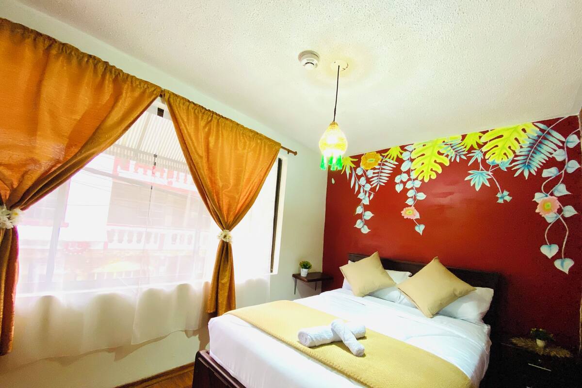 Habitación de hotel con cama, mesita de noche, ventana con cortinas y un mural floral en la pared.