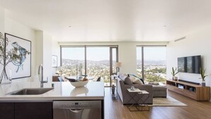 A modern kitchen with a dishwasher, a dining area with a bowl of fruit, a sofa, and a TV. - Visit LA in Ultra Luxury with Breathtaking Views (Beverly Hills)