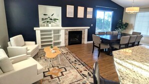 A living room with a fireplace, white sofas, a dining table with chairs, and a kitchen area with a granite countertop.