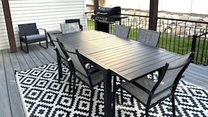 An outdoor patio with a wooden table and chairs, a grill, and a patterned rug.