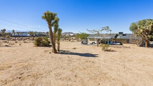 A desert landscape with sparse vegetation, including a prominent Joshua tree, and a building with a sloped roof. - Starlight Ranch with Hot tub, Fire pit & BBQ! (Joshua Tree)