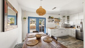 A modern kitchen with white cabinets, a wooden dining table with woven placemats, and a hanging rattan light fixture. - Starlight Ranch with Hot Tub, Fire Pit & BBQ! (Joshua Tree)