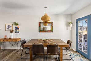 A dining area with a wooden table, four chairs, a hanging woven light fixture, and a colorful mirror on the wall.