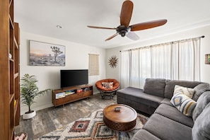 A living room with a sofa, a television, a ceiling fan, and a large window with curtains.