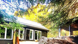 Une cabane avec un escalier en pierre menant à une terrasse en bois, entourée d’arbres et d’un grand rocher.