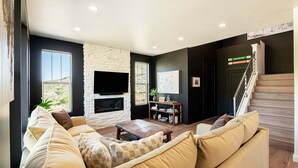 A living room with a stone fireplace, a flat-screen TV, a staircase, and a view of the outdoors. - Powder and Lake Days Await | Luxury and Comfort w/ Views & Private Hot Tub (Heber City)