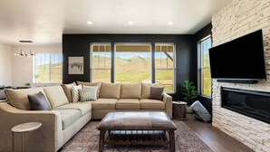 A living room with a sectional sofa, a coffee table, a fireplace, and large windows with a view of hills. - Powder and Lake Days Await | Luxury and Comfort w/ Views & Private Hot Tub (Heber City)