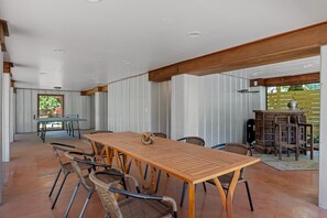 Een ruime patio met een lange houten tafel, rieten stoelen, een klein barretje en uitzicht op groen.