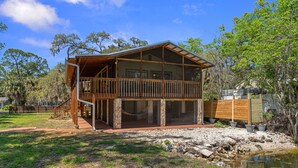 A two-story wooden cabin with a balcony, a stone foundation, and a pond in the foreground. - Peaceful Waterfront Escape on Withlacoochee • Private Dock, Old Florida Charm (Yankeetown)
