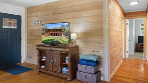 A room with wooden panel walls, a television on a wooden cabinet, a door with glass panels, and a small table with a lamp. - Peaceful Waterfront Escape on Withlacoochee • Private Dock, Old Florida Charm (Yankeetown)