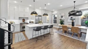A modern kitchen with white cabinets, a central island with bar stools, pendant lighting, and a dining area with a wooden table and chairs. - Uptown Brownstone - 1 Block off Downtown Square (Bentonville)