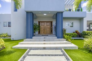Un bâtiment moderne à la façade symétrique, doté d’une entrée centrale avec une double porte en bois, flanquée de piliers bleus, et d’un petit escalier menant à l’entrée.
