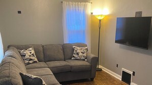 Ein Wohnzimmer mit grauem Ecksofa, Deckenventilator, Stehlampe und einem an der Wand befestigten Flachbildfernseher.