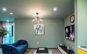 A modern living room with a blue sofa, a contemporary light fixture, a framed abstract painting, a clock, and a digital display on the wall.