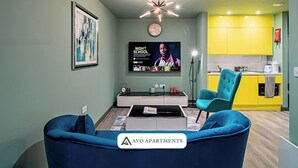 Een modern appartement met een woonkamer, inclusief een blauwe bank, een salontafel en een tv waarop 'Night School' te zien is. De keuken heeft gele kastjes en een spoelbak.