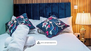 Hotelkamer met een wand van houten panelen, een donkerblauwe hoofdbord, twee decoratieve kussens met bloemmotieven, een witte spreidek en een witte badjas, plus een bedlampje.