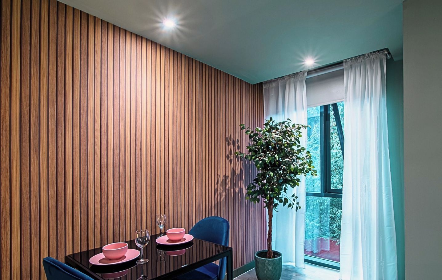 A modern dining area with a wooden panel wall, a dining table set with pink plates and glasses, blue chairs, and a potted plant.