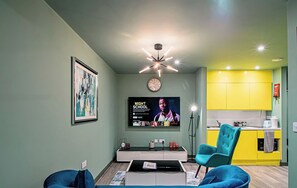 A modern apartment with a living room featuring a blue sofa, a coffee table, and a TV displaying 'Night School'. The kitchen has yellow cabinets and a sink. - Studio in Sheffield City Centre (South Yorkshire)