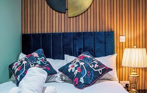 Une chambre d’hôtel avec un mur recouvert de panneaux de bois, une tête de lit bleu foncé, deux coussins décoratifs à motifs floraux, un couvre-lit blanc, un peignoir blanc et une lampe de chevet.