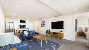 A modern living room with a sofa, coffee table, and TV. There is a kitchen area in the background. - Unit 2907 Oracle Tower 1 Level 29 (Broadbeach)