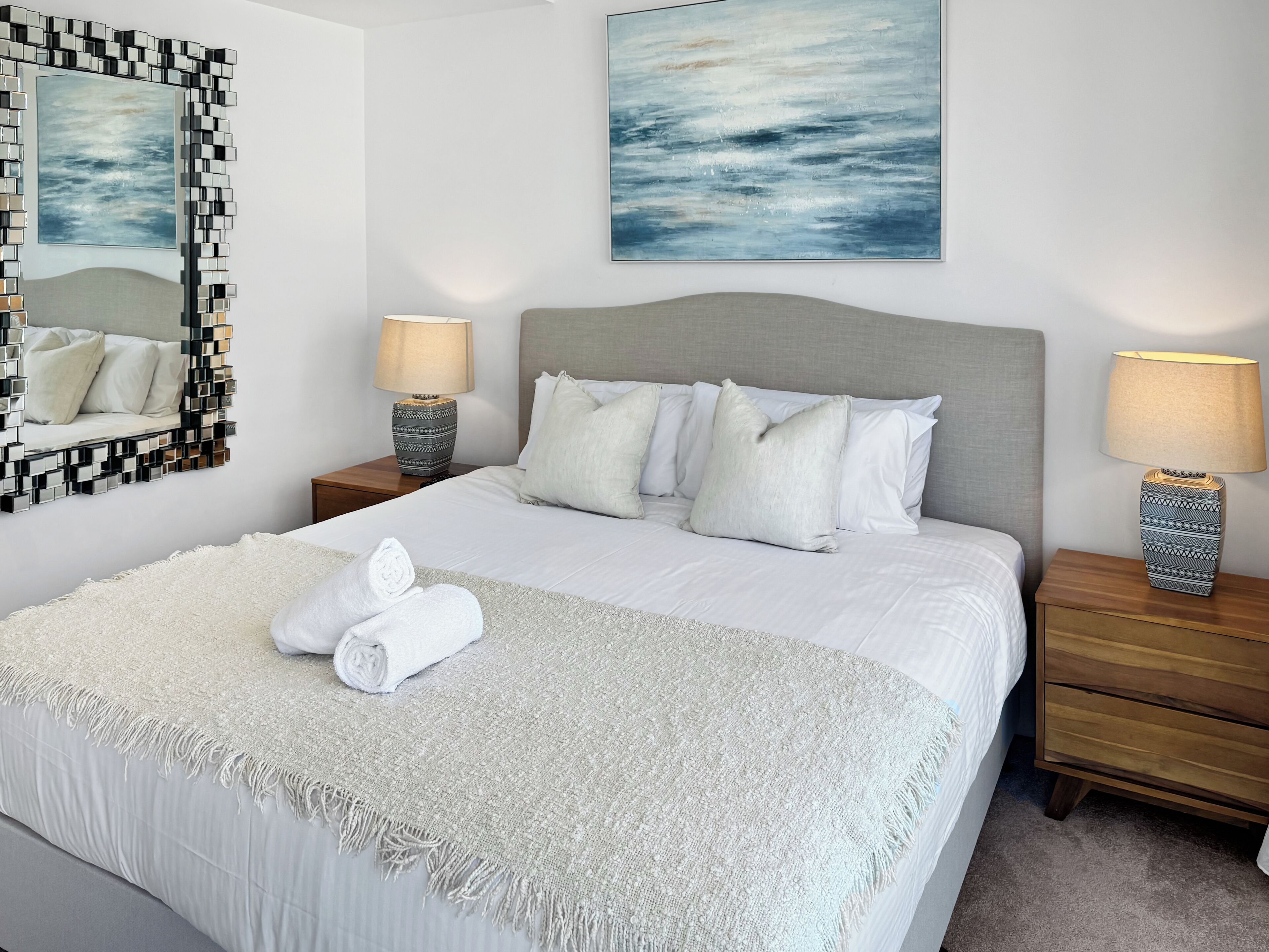 A neatly made bed with white linens, a grey headboard, and a white throw blanket. There are two bedside tables with lamps and a large abstract painting above the bed.
