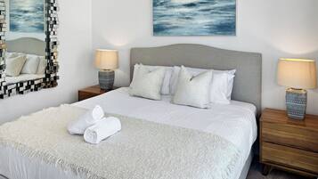 A neatly made bed with white linens, a grey headboard, and a white throw blanket. There are two bedside tables with lamps and a large abstract painting above the bed.
