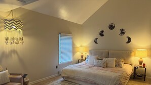 Une chambre à coucher avec un grand lit, deux tables de chevet avec des lampes, un tapis à motifs, une chaise et des éléments de décoration murale, notamment des croissants de lune.